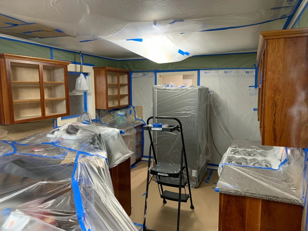 Kitchen sealed in plastic sheeting, blue tape and green paper, with a step ladder, before spraying