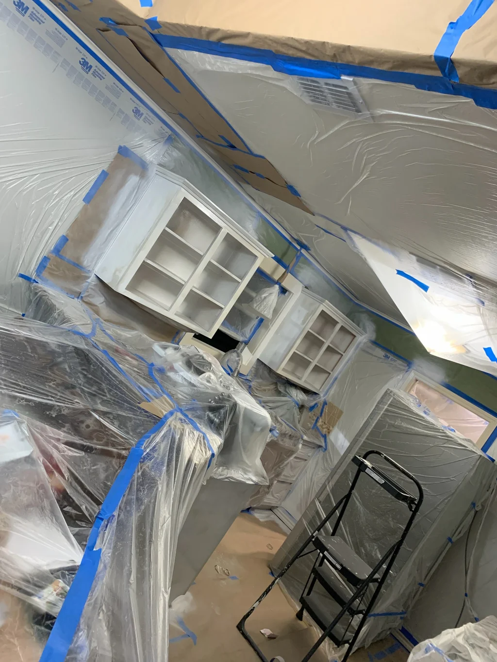 Glass front cabinets freshly sprayed white inside the plastic containment, tape still on the edges