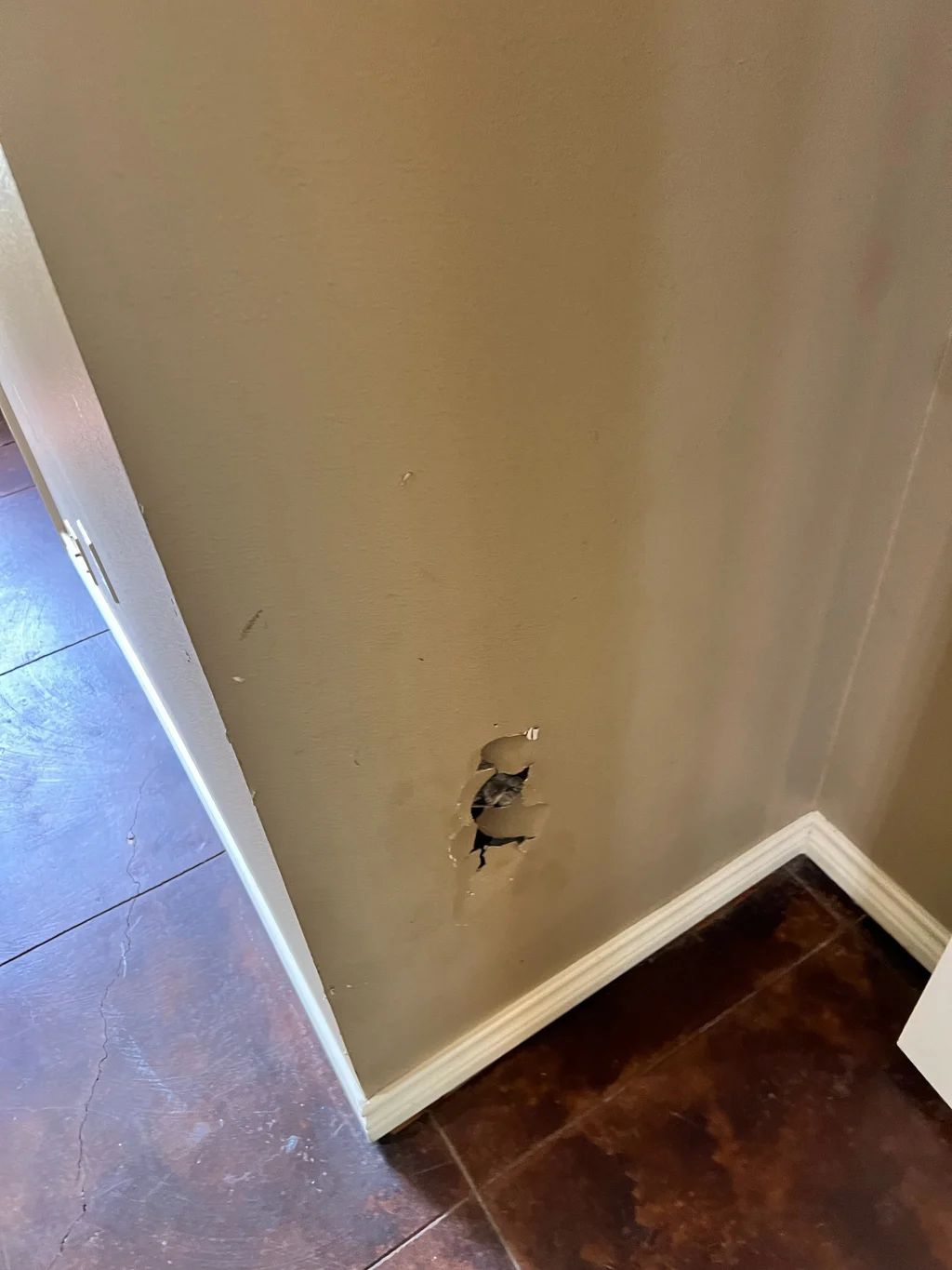 Hole punched through drywall above the baseboard in a tan hallway, before any repair
