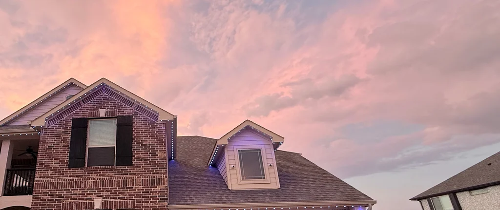 Brick two story home at sunset with rooflines and a dormer lit in violet
