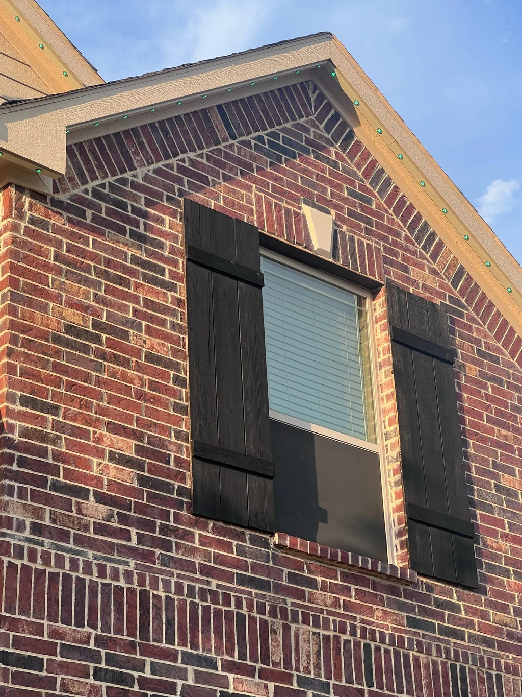 Lighting track on a red brick gable rake beside a lower adjoining roofline, shuttered window below